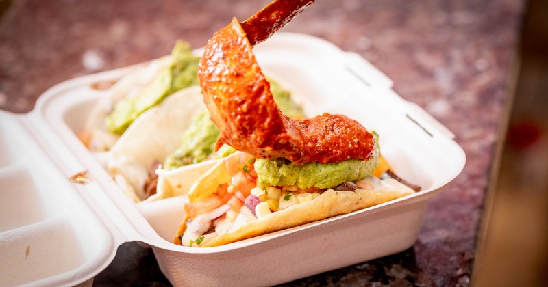 Spoon adding red salsa to tacos topped with guacamole in a takeout container