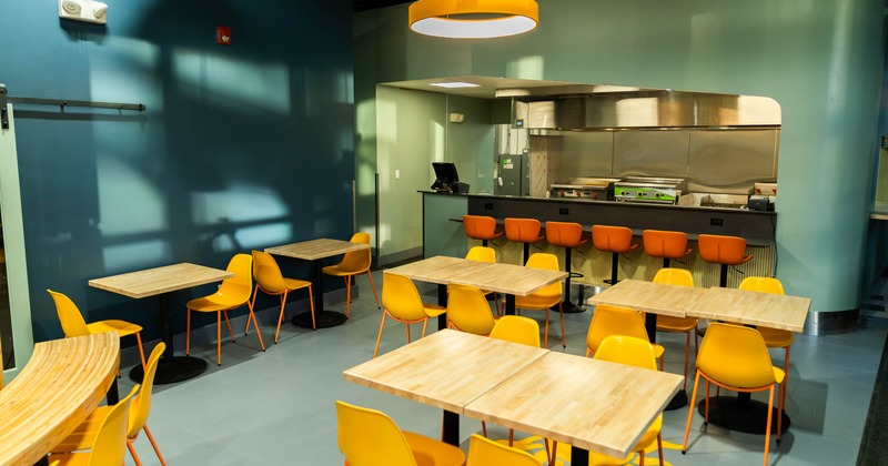 A modern dining area with wooden tables, yellow chairs, and a kitchen in the background