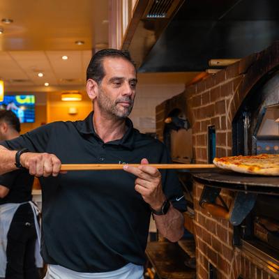 Pizza maker uses a wooden peel to remove a fresh pizza from a brick oven in a kitchen.