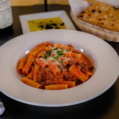 Bowl of Rigatoni Bolognese served alongside focaccia, olive oil and a glass of red wine.