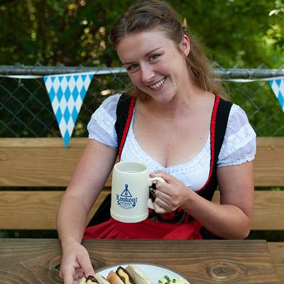 Person in Oktoberfest-style outfit holding a beer mug at an outdoor table.