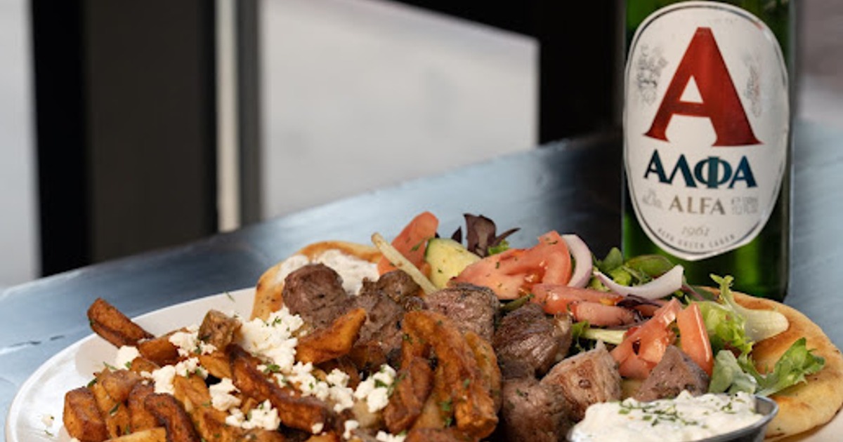 A plate of Greek food with skewered meat next to a bottle of beer on a table