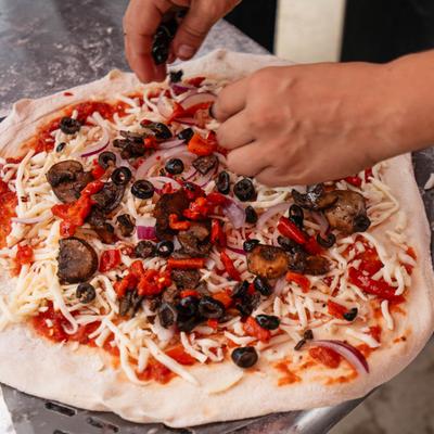 Hands topping a veggie pizza with mushrooms, black olives, and red peppers.