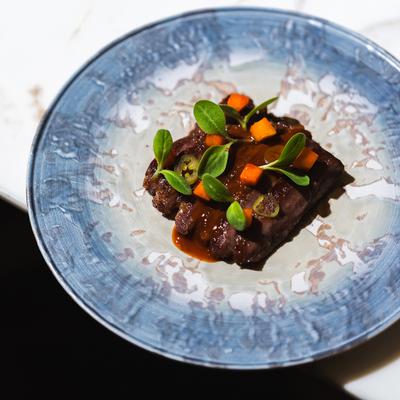 Pork cheek dish on a textured blue plate, on a light marble surface.