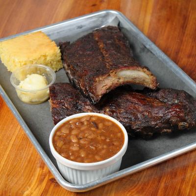 Ribs served with cornbread, a small cup with butter, and a side of baked beans.