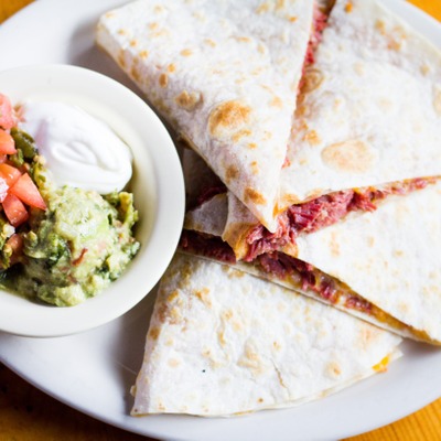 Quesadilla stuffed with corned beef on a plate served with a dish that contains guacamole, sour cream and pico de gallo