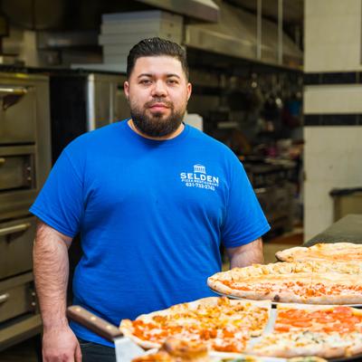Chef  in blue shirt behind trays of pizza in a kitchen setting.