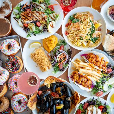 Overhead view of a wooden table laden with diverse dishes, coffees, and cocktails.