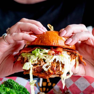 Hands holding a pulled pork sandwich with bacon and coleslaw.