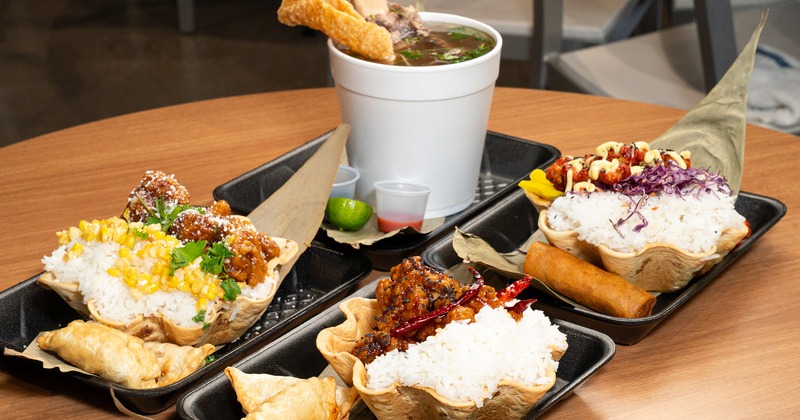 Assorted Asian-inspired bowls with rice, chicken, corn, soup, and spring rolls on a wooden table
