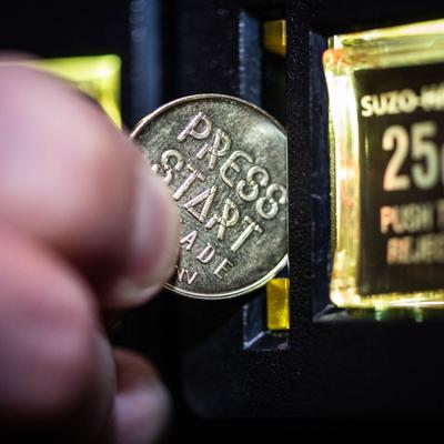 Close-up of a hand inserting a coin into an arcade machine slot, the coin reads PRESS START.