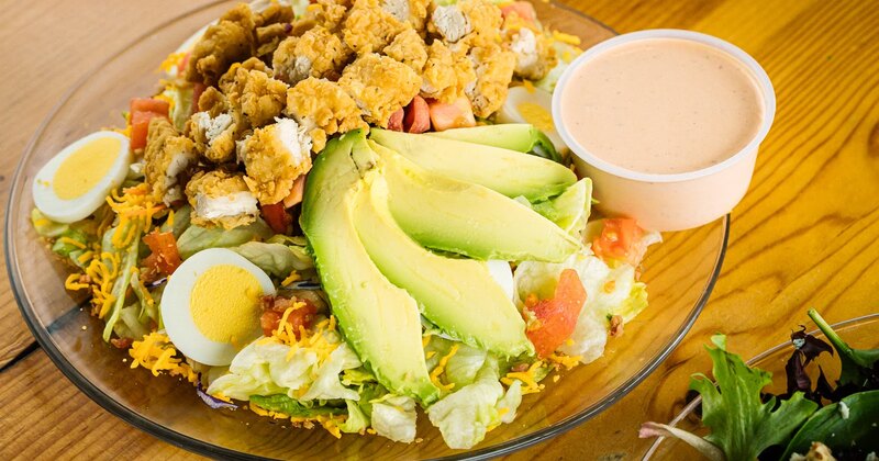 Salad with fried chicken, avocado, hard-boiled egg, cheese, bacon, and a cup of dressing.