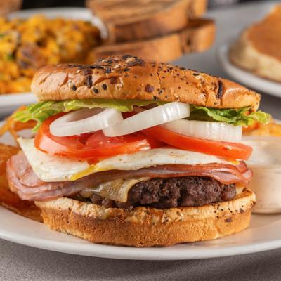 Burger on a seeded bun topped with ham, fried egg, tomatoes, and onions.