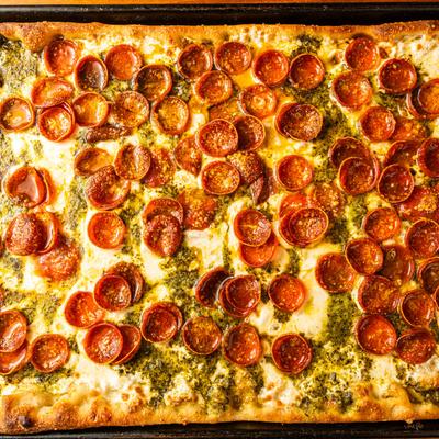 Rectangular pizza topped with melted cheese, green pesto, and pepperoni.