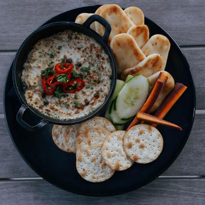 A black plate with white fish dip, pita, crackers, cucumber, and carrot sticks.