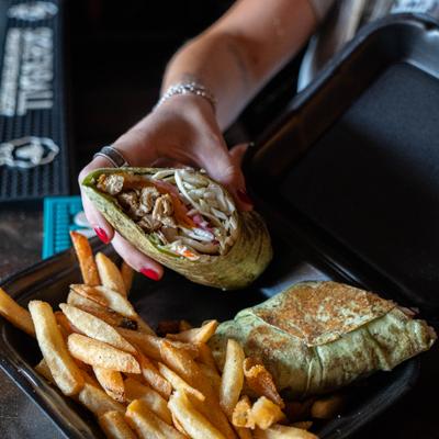 Hand grabbing a chicken wrap half from a takeout container filled with fries.