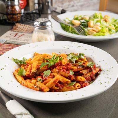 Penne Arrabbiata plate sits on the table in the foreground, with Caesar salad behind it.