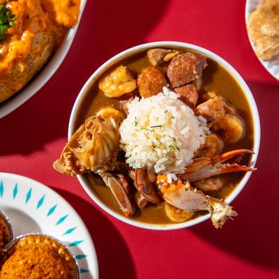 A bowl of seafood gumbo sits on a red table with other dishes.