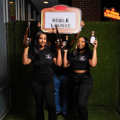 Three people posing with bottles and a sign.