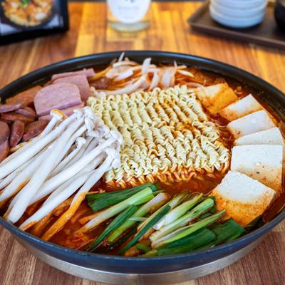 Spicy ramen stew with noodles, tofu, mushrooms, ham, sausage, and green onions in a black pot.