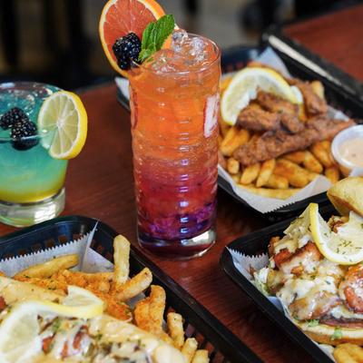 Two colorful cocktails with fruit garnishes surrounded by comfort food.