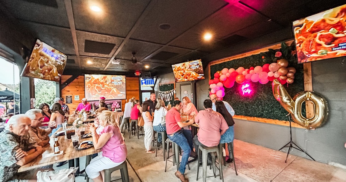 A party with guests at tables in front of a green leaves backdrop with pink balloons