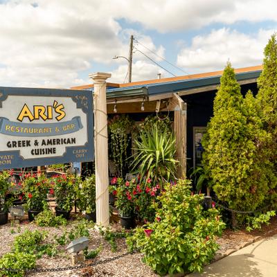 The exterior signage for Ari's Restaurant & Bar, surrounded by lush greenery and colorful flowers.