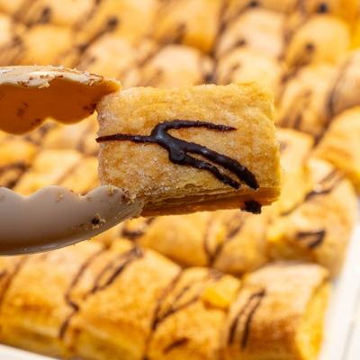 Tongs hold a square, chocolate-drizzled pastry above rows of similar golden baked goods.