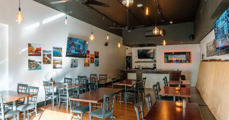 Bright café interior with wood tables, gray chairs, hanging Edison bulbs, wall photographs, televisions, and a tiled bar