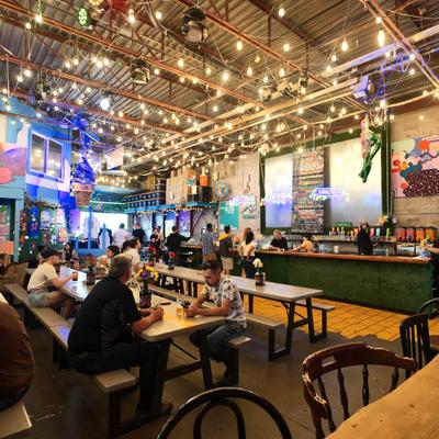 People enjoying drinks in an indoor beer hall with long tables and string lights.