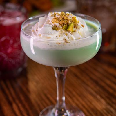A green dessert cocktail topped with cream and pistachio in a stemmed glass.