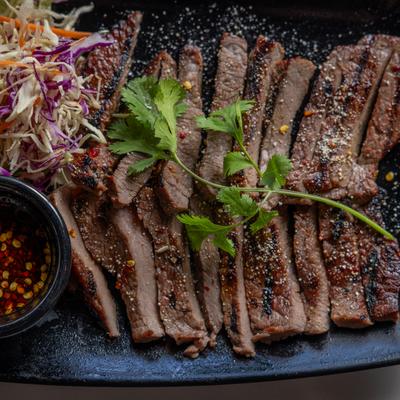 Grilled beef slices on a black plate, accompanied by slaw and a small bowl of chili sauce.