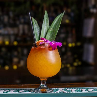 An orange cocktail, garnished with pineapple leaves, purple flower and dry lemon wheel.