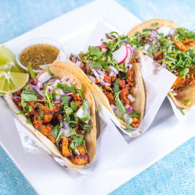 Three chicken tacos topped with cilantro and onions, served with green salsa and lime wedges.