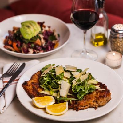 Breaded pork chop with arugula, Parmesan, lemon wedges, and a glass of red wine.