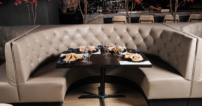 Circular booth with beige leather seating, set with glassware and napkins, beneath artificial trees