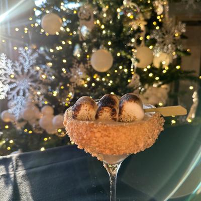 Cocktail drink garnished with toasted marshmallows sits on a table, Christmas tree behind it.