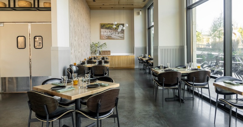 Restaurant interior with wooden tables, black chairs, pendant lights, and large windows