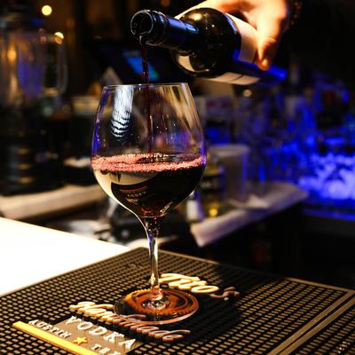 A bartender pours red wine into a wine glass on a bar counter with dim lighting.
