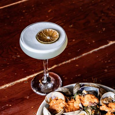 Sage Gimlet cocktail garnished with dehydrated lemon wheel, a plate with clams.