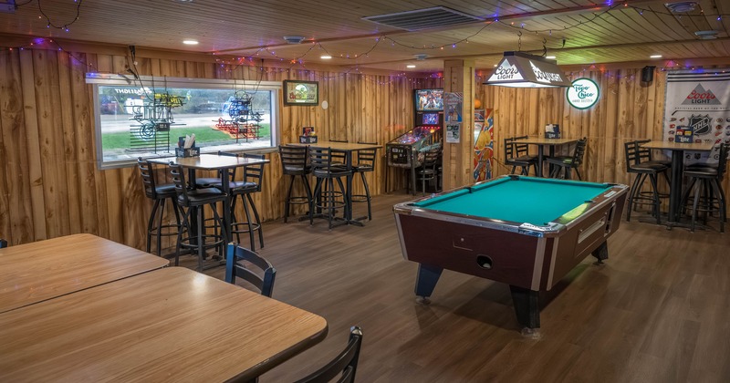 Interior of a rustic recreational room with a pool table, bar stools, pinball machine