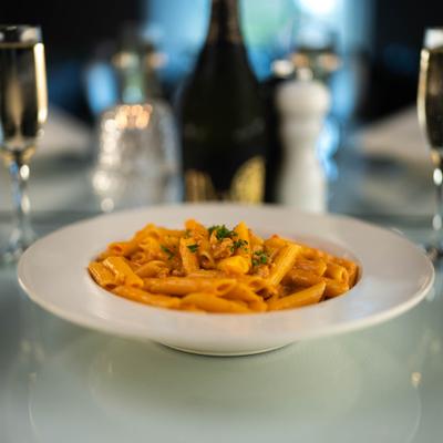 Penne alla Lenzi plate on a white table, flanked by two flutes of sparkling wine.