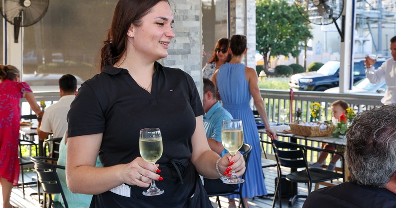 Server bringing two glasses of white wine to guests seated on a covered patio