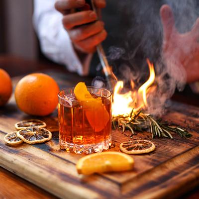 Bartender smoking a sprig of rosemary on a wooden board, alongside a cocktail.