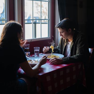 A couple eating pasta at a checkered tablecloth restaurant table near a window.