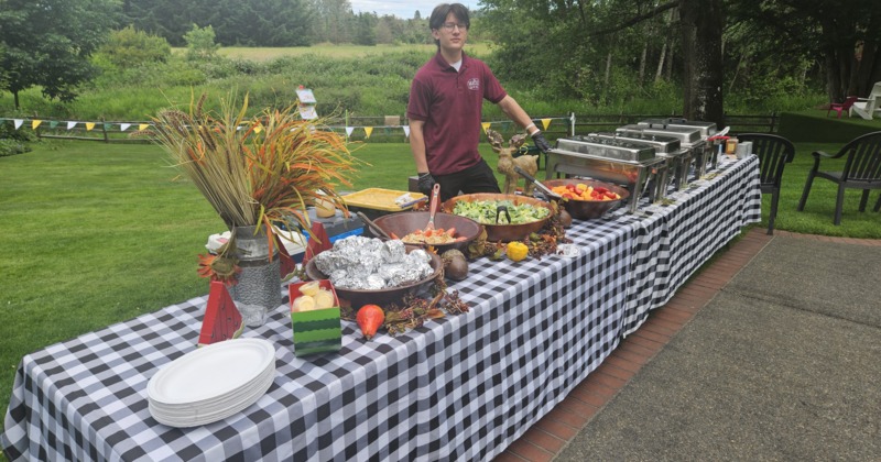 Outdoor buffet table with assorted dishes and a server on a grassy lawn