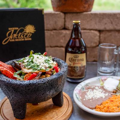 Molcajete De Mariscos sits next to a plate of  rice and beans, a beer bottle, and mugs.