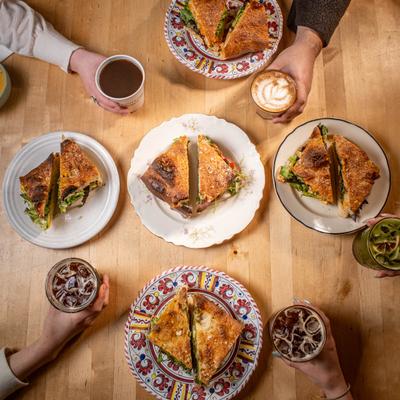 People holding iced drinks around toasted sandwiches on a wooden table, overhead view.