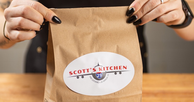 Hands holding a brown paper bag with a white sticker displaying Scott's Kitchen logo