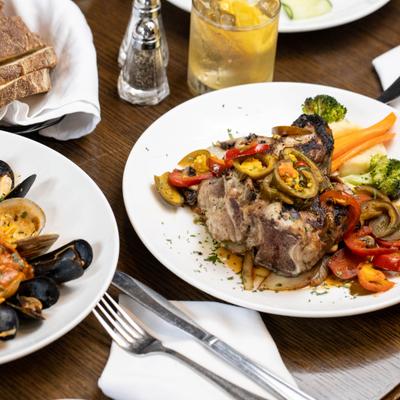Close up of a table with seafood pasta and a veal chop dish, accompanied with bread and a drink.